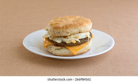 A Sausage Egg And Cheese Biscuit Breakfast Sandwich On A White Plate Upon A Tan Table Cloth.