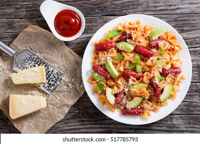Sausages Bowtie Pasta Warm Salad With Avocado Slices And Basil Leaves, Sprinkle With Cheese On White Plate On Wooden Table With Grater On Piece Of Paper With Grated Parmesan Cheese,  View From Above