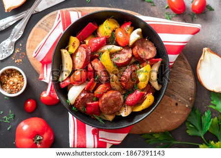 Sausages fried with peppers, tomatoes, and onions in a black plate on a dark background top view. Sausages stewed with vegetables. Traditional Hungarian food lecho.