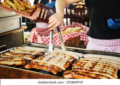 Sausages on the grill, hands of the seller and a pile of  buns at background. Hot dogs stall at  famous Borough Food Market (London, England). Selective focus on the sausages at the left corner.