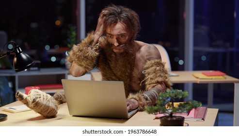 Savage office worker in prehistoric outfit sitting on office desk opening laptop getting scared starting interacting with modern technology device. Nighttime. Humor concept,