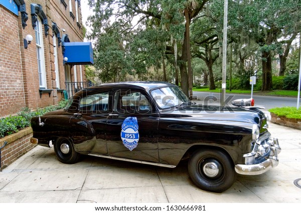 Savannah, Georgia / USA - August 18 2012:  Black\
vintage police car from 1953, in front of headquarters of the\
Savannah-Chatham Police Department, a brick building with a blue\
canopy and trees\
behind