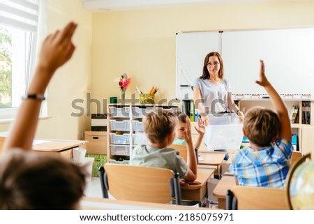 Save the earth. Charming female teacher with small school children in classroom learning about waste separation. Children raised their hands to answer, rear view.
