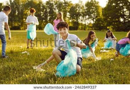 Save planet. Portrait of little boy who collects trash in garbage bag in forest or in park. Smiling preteen boy collects trash on green lawn in background of his peers who pick up used plastic.