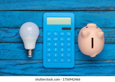 Saving Electricity. Led Light Bulb And Piggy Bank, Calculator On Blue Wooden Background. Top View