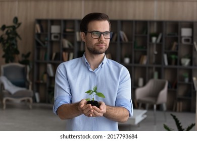 Saving environment. Peaceful businessman in eyewear stand at home in office hold small green plant in palms look at distance. Young male entrepreneur take care of nature think on ecological production