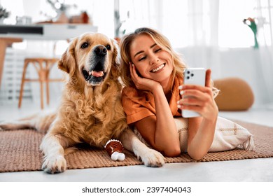 Saving memories with pet. Smiling woman with blond hair snuggling to furry friend and taking selfie on modern cell phone. Obedient golden retriever lying on floor near delighted female owner. - Powered by Shutterstock