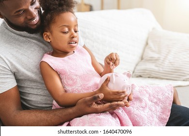 Saving Money Concept. Close Up Of Handsome African Father Showing Cute Little Daughter Piggy Bank, Home Interior, Copy Space