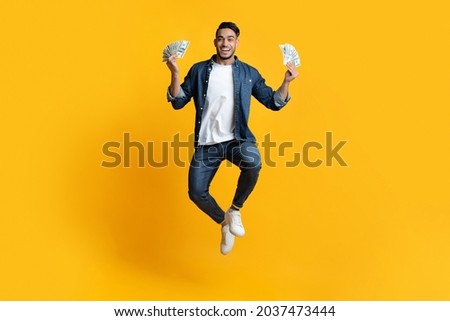 Saving money, gambling, gaming, trading, loan concept. Delighted middle-eastern guy holding bunch of money in both hands and smiling, won lottery, yellow studio background, full length, copy space