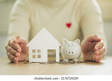 Saving Money For A House And Real Estate. Women's Hands Protect The Piggybank And House Shape In Hand, Family Money Management, Family Finance Plan Concept, Saving Money, And Investment Concept. 