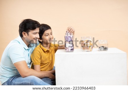 Saving Money and Investing Concept Happy Young Indian Father and Son Put Rupee Notes Or Cash In Multiple Glass Jar Bottles Isolated On Beige Studio Background.