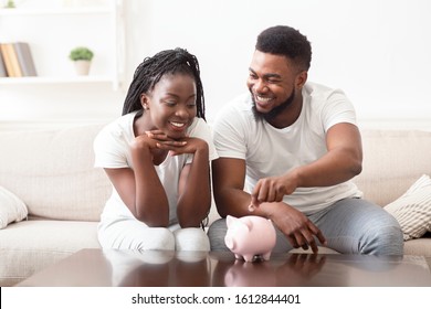 Savings For Future. Young Black Couple Putting Coin In Piggybank, Adding Money To Family Budget, Sitting Together On Sofa At Home