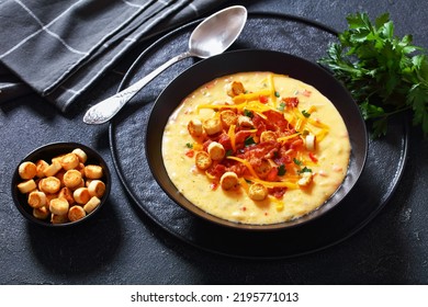 Savory Creamy Pumpkin Soup Topped With Fried Bacon, Shredded Cheddar Cheese And Salty Croutons In Black Bowl On Concrete Table With Spoon