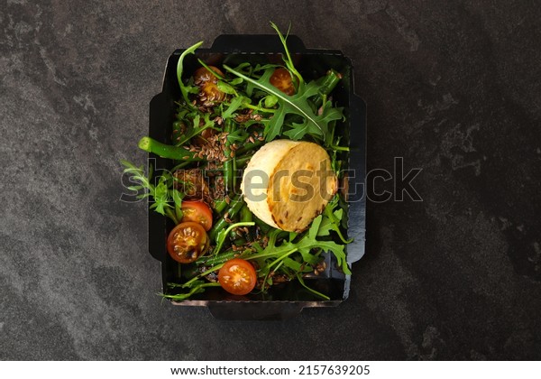 Savory souffle salmon and salad with arugula,\
tomatoes and green beans in black paper takeaway box on dark\
background. Healthy food delivery\
concept.