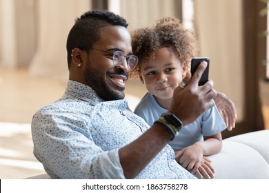 Say cheese, sonny! Happy black dad sitting on couch in living room cuddling cute little boy and taking selfie on smartphone, smiling african father showing son kid funny video or cartoon on cellphone