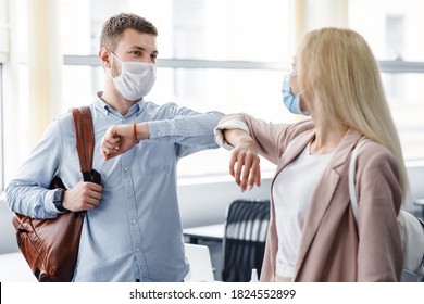 Say Hello And Greet, Social Distance And Return To Work After Quarantine. Millennial Man And Woman In Protective Masks Are Touched By Elbows In Interior Of Office During COVID-19 Epidemic, Free Space