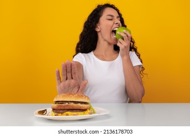 Say No To Junk Food. Lady Refusing To Eat Unhealthy Burger Choosing Biting Apple, Showing Stop Sign Gesture With Palm Hand To Hamburger Sitting At Table, Selective Focus, Yellow Orange Studio Wall