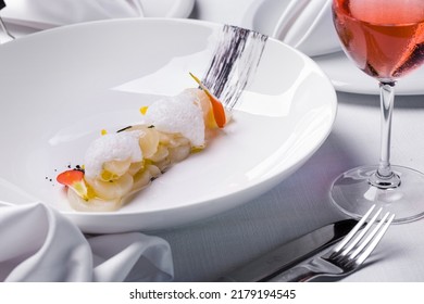 Scallop, Finely Chopped, Stacked In A Slide, Top With Citrus Foam And Edible Petals. The Food Is In A Deep White Plate. The Plate Is On A White Tablecloth, Next To It Are Cutlery And Cloth Napkins, An