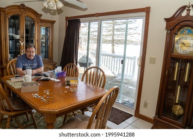 Scandia, Minnesota - Jan 25, 2021: Senior Mature Man Sits At A Messy Kitchen Table, Playing With IPad Tablet As A Puzzle Is Strewn About On The Table