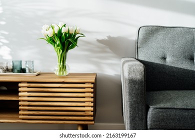 Scandinavian home interior with spring bouquet of white tulip flowers in glass vase, tray with candles standing on a wooden cabinet. Minimalist design with gray sofa and white wall. Springtime.