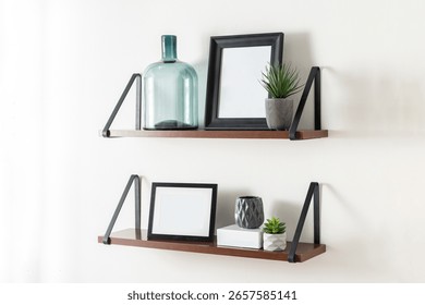 Scandinavian inspired composition featuring two floating wooden shelves with black metal brackets, decorated with framed pictures, green glass bottle, mini succulents and ceramic vases on white wall.