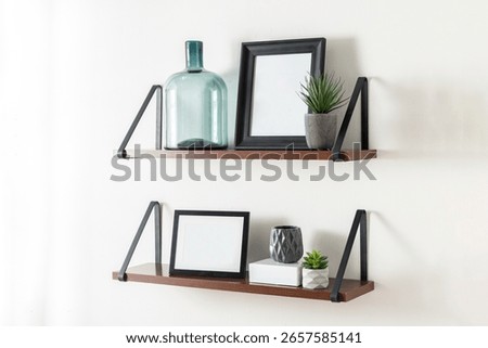 Scandinavian inspired composition featuring two floating wooden shelves with black metal brackets, decorated with framed pictures, green glass bottle, mini succulents and ceramic vases on white wall.