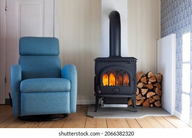 Scandinavian interior: wood burning stove, comfy blue recliner armchair, firewood in corner and sunlight on the wall. 