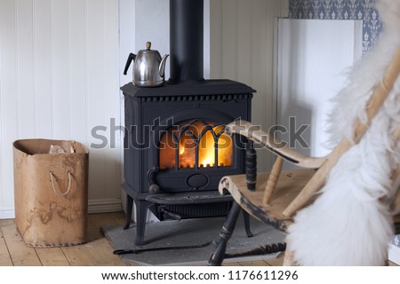 Scandinavian interior: wood burning stove, box of firewood and old restored rocking chair in living room corner. Metal coffeepot at stove. White sheep skin in rocking chair.
