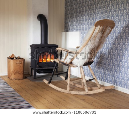 Scandinavian interior: wood burning stove, box of firewood and old restored rocking chair in living room corner. Rag carpet on oiled wooden floor. White sheep skin in rocking chair. 
