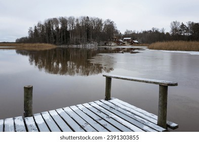 Scandinavian landscape with a lake in late autumn, Small wooden bridge sprinkled with fresh snow, Traditional Swedish red houses and a forest with bare trees reflected in calm water of Lake Mälaren - Powered by Shutterstock