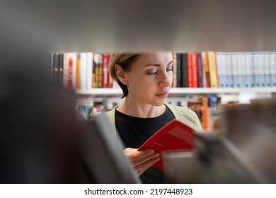 Scandinavian Middle-aged Woman Looking For Book At Library Bookshelf. Female Mature Student Searching For Novel Or Textbook At Bookshelf, Studying At University Or College Campus Building