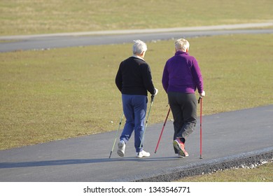 Scandinavian Nordic Walking. Two Older Women Are Engaged In Sports Even Down The Paved Path In The Spring Time. In The Rays Of Sunlight. Healthy Lifestyle. Sport's Event. A Example Of Society.