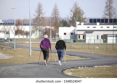 Scandinavian Nordic Walking. Two Older Women Are Engaged In Sports Even Down The Paved Path In The Spring Time. In The Rays Of Sunlight. Healthy Lifestyle. Sport's Event. A Example Of Society.