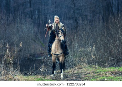 Scandinavian northern viking riding horse with ax in hand. Northern warrior woman riding in forest in war clothes with fur collar, war makeup.