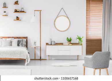 Scandinavian Style White Room Interior With Round Mirror On The Wall, Wooden Bed With Pillows And Blanket, Cabinet And Armchair. Real Photo.