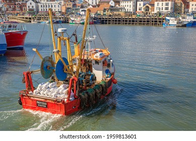 Scarborough, UK.  April 16, 2021.  A Fishing Trawler Enters A Harbour. A Waterfront Is Visible Ahead And Wake Trails Behind.  White Sacks Are Stacked At The Stern.