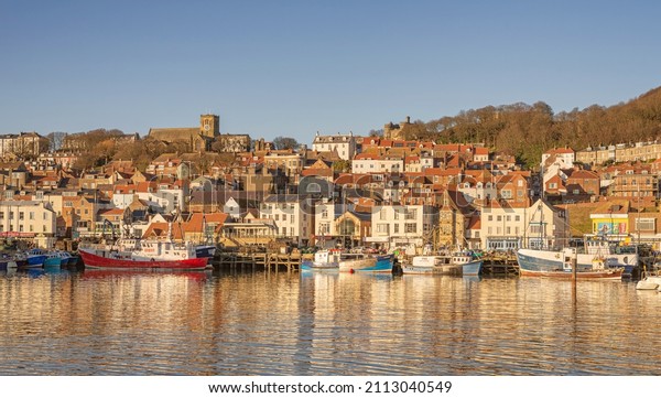 Scarborough, UK. January
13, 2022. A low winter sun falls on the buildings of a town set on
a hillside. The harbour below has trawlers  moored to a quay and a
clear sky is
above.