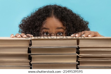 Scared African American female student hiding behind stacks of books, being afraid of college exam on blue studio background. Frightened black lady with textbooks studying for university test