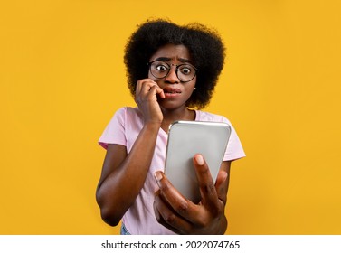 Scared african american woman holding mobile phone seeing bad news, photos or message and looking at camera over yellow background. Human reaction, expression