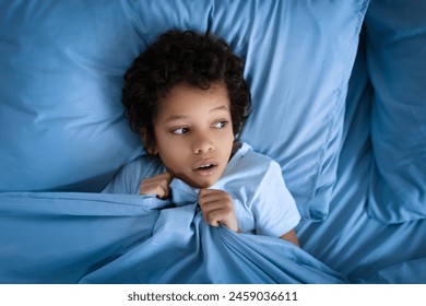 Scared African American young boy with curly hair lies awake in a blue bed during night, clutching the bedding close to his chin with a look of fright - Powered by Shutterstock