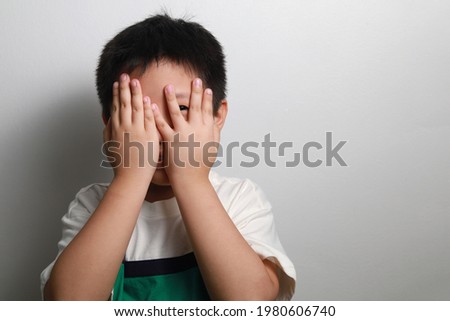 A scared Asian boy peeks through his fingers, standing against a grey wall.
