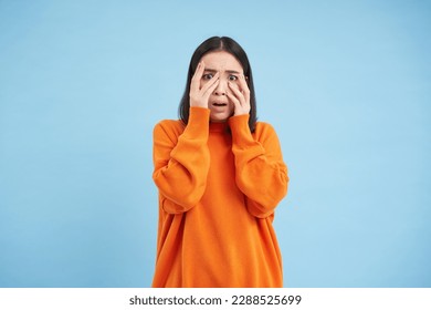 Scared Asian woman shuts her eyes and peeks through hands at smth scary and disturbing, shocked by smth horrifying, standing over blue background.