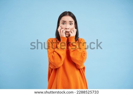 Scared Asian woman shuts her eyes and peeks through hands at smth scary and disturbing, shocked by smth horrifying, standing over blue background.