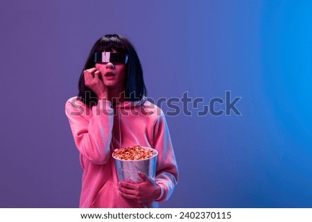 Scared awesome brunet woman in pink hoodie trendy specular sunglasses with popcorn hold hand on cheek posing isolated in blue violet color light background. Neon party Cinema concept. Copy space