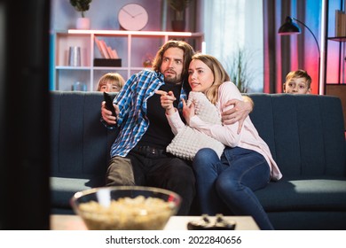 Scared Bearded Husband Hugging His Frightened Wife During, Watching Horror Film On TV At Home In The Evening. Two Scared Kids Are Hidden From Scare Behind The Sofa