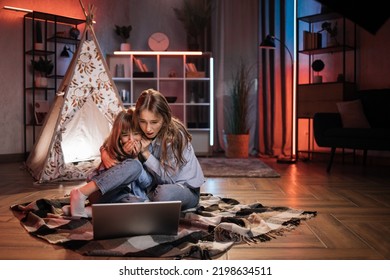 Scared Blond Woman, Nanny Or Older Sister Hugging While Watching Horror Movie With Her Lovely Younger Sister Outside Teepee Tent. Evening Leisure And Family Concept.