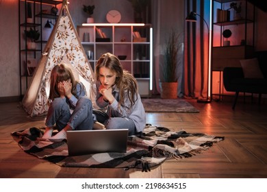 Scared Blond Woman, Nanny Or Older Sister Hugging While Watching Horror Movie With Her Lovely Younger Sister Outside Teepee Tent. Evening Leisure And Family Concept.