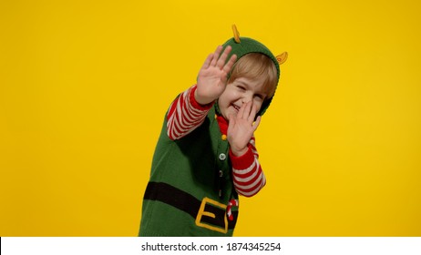 Scared Blonde Girl In Christmas Elf Santa Helper Costume Looking At Camera Afraid Cover Hiding Face With Hands. Child Covering Face With Fearful Expression Hides From Something Terrifying