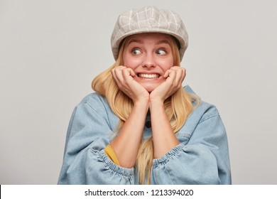 Scared blonde girl looks aside frightened afraid chattering teeth with fear,bites nails from fright, hands holding cheeks, wears oversize denim jacket and beige checked cap,on grey background