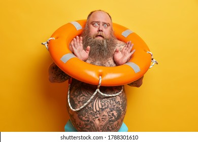 Scared Bugged Eyed Man With Thick Beard And Tattooed Body, Afraids Of Swimming, Carries Inflated Lifebuoy, Isolated On Yellow Background. Overweight Guy Spends Summer Time On Beach. Lifestyle Concept